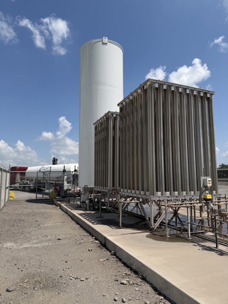 A tall white industrial storage tank stands beside a corrugated metal processing unit, with pipes and equipment on a concrete pad under a blue sky.
