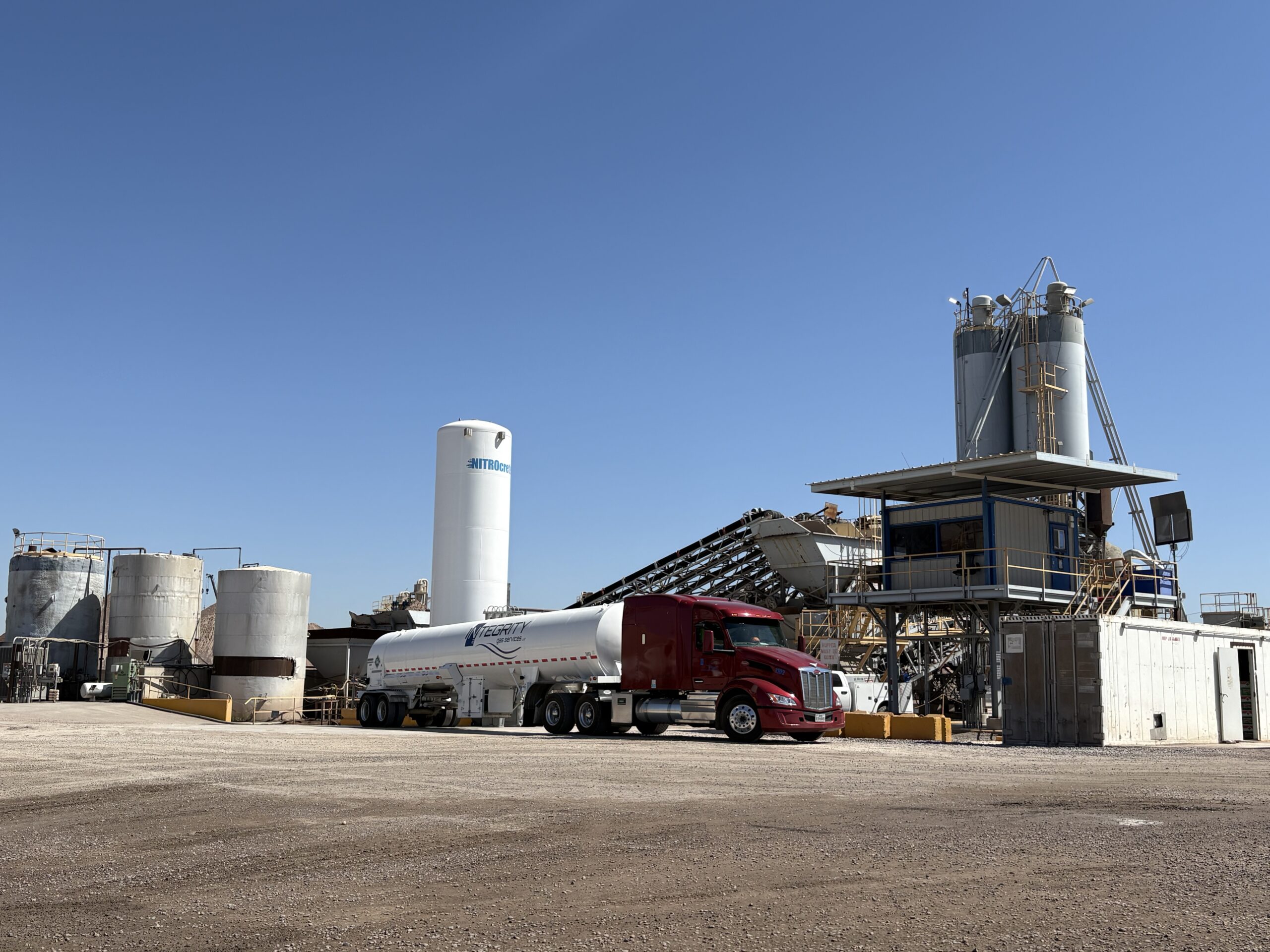 A red tanker truck is parked at an industrial plant with large storage tanks, silos, and conveyor structures under a clear blue sky.