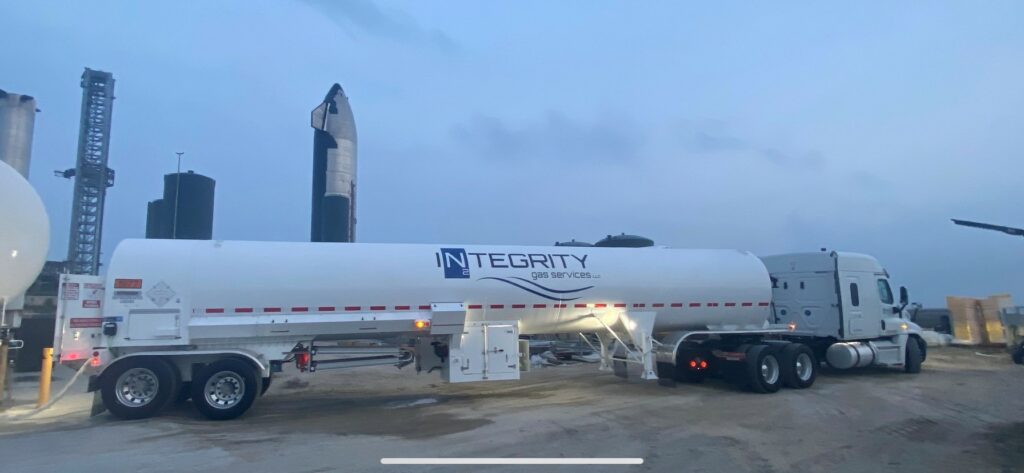 A white Integrity tanker truck parked at an industrial site at dusk, with towers and equipment in the background.